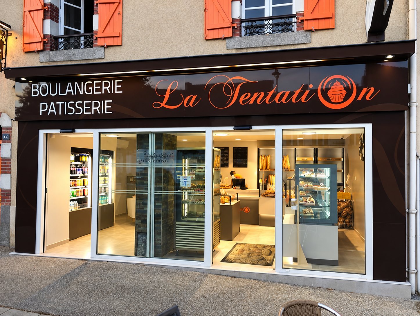 Façade de la boulangerie-pâtisserie La Tentation, avec son enseigne brune, ses grandes vitrines éclairées et ses volets orange.