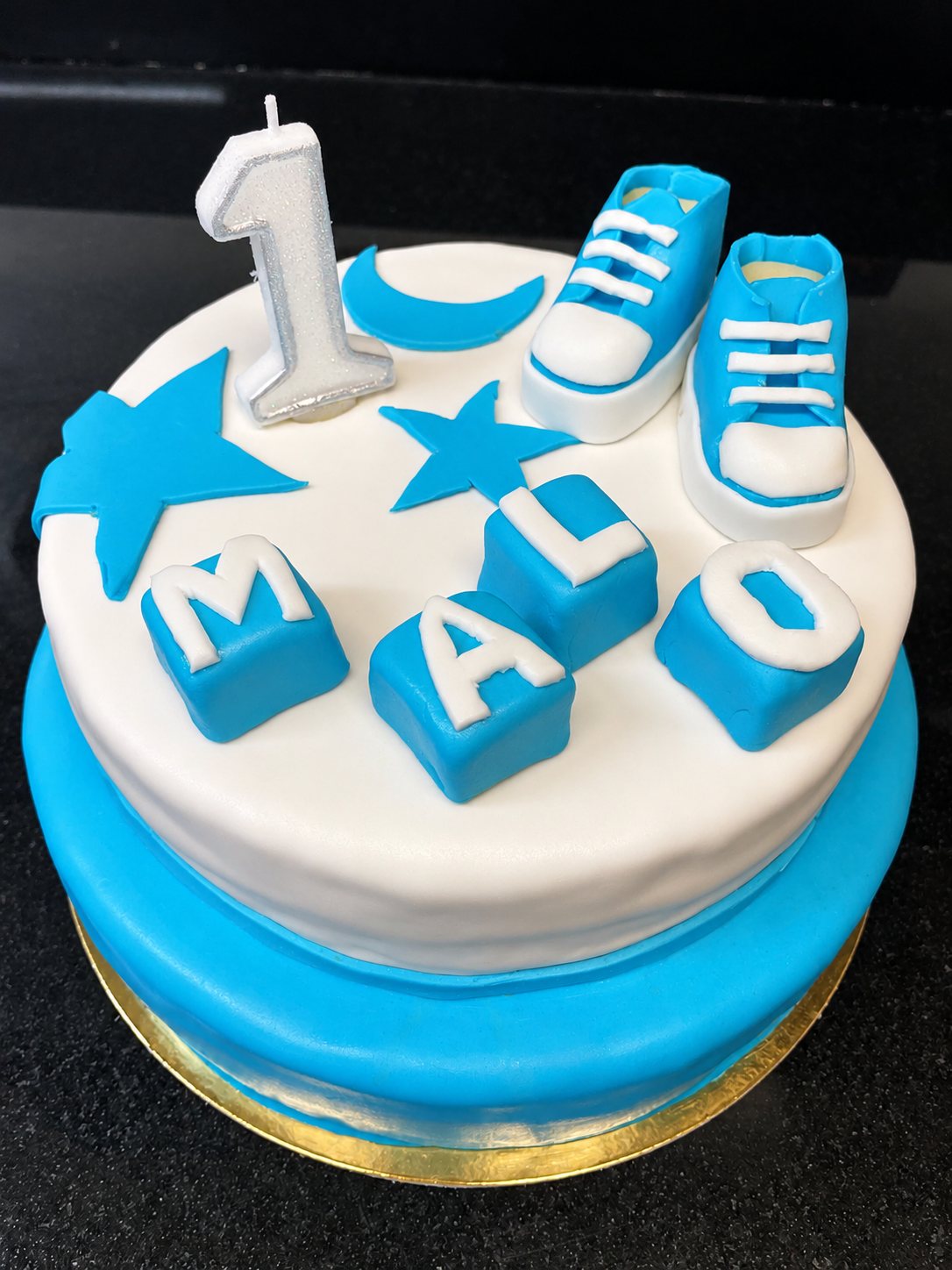 Gâteau d'anniversaire en pâte à sucre bleu et blanc, décoré d'une bougie en forme de 1, de petites baskets et de cubes lettrés.