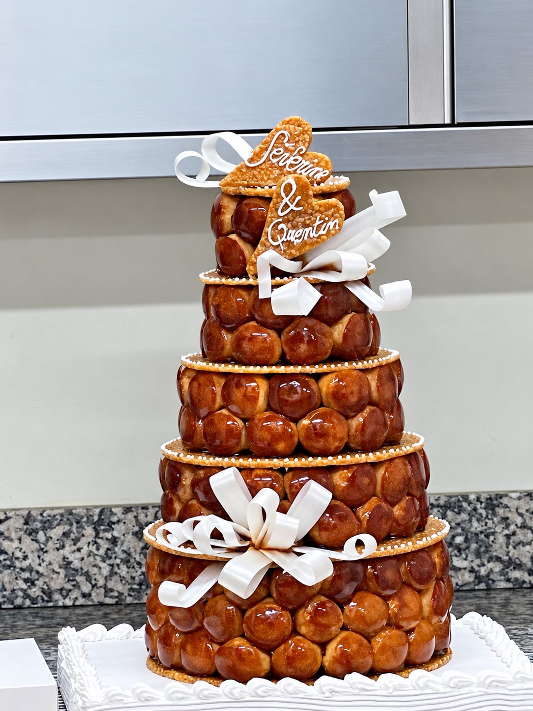 Pièce montée de choux caramélisés à cinq étages, décorée de rubans blancs et de deux cœurs en pâte.