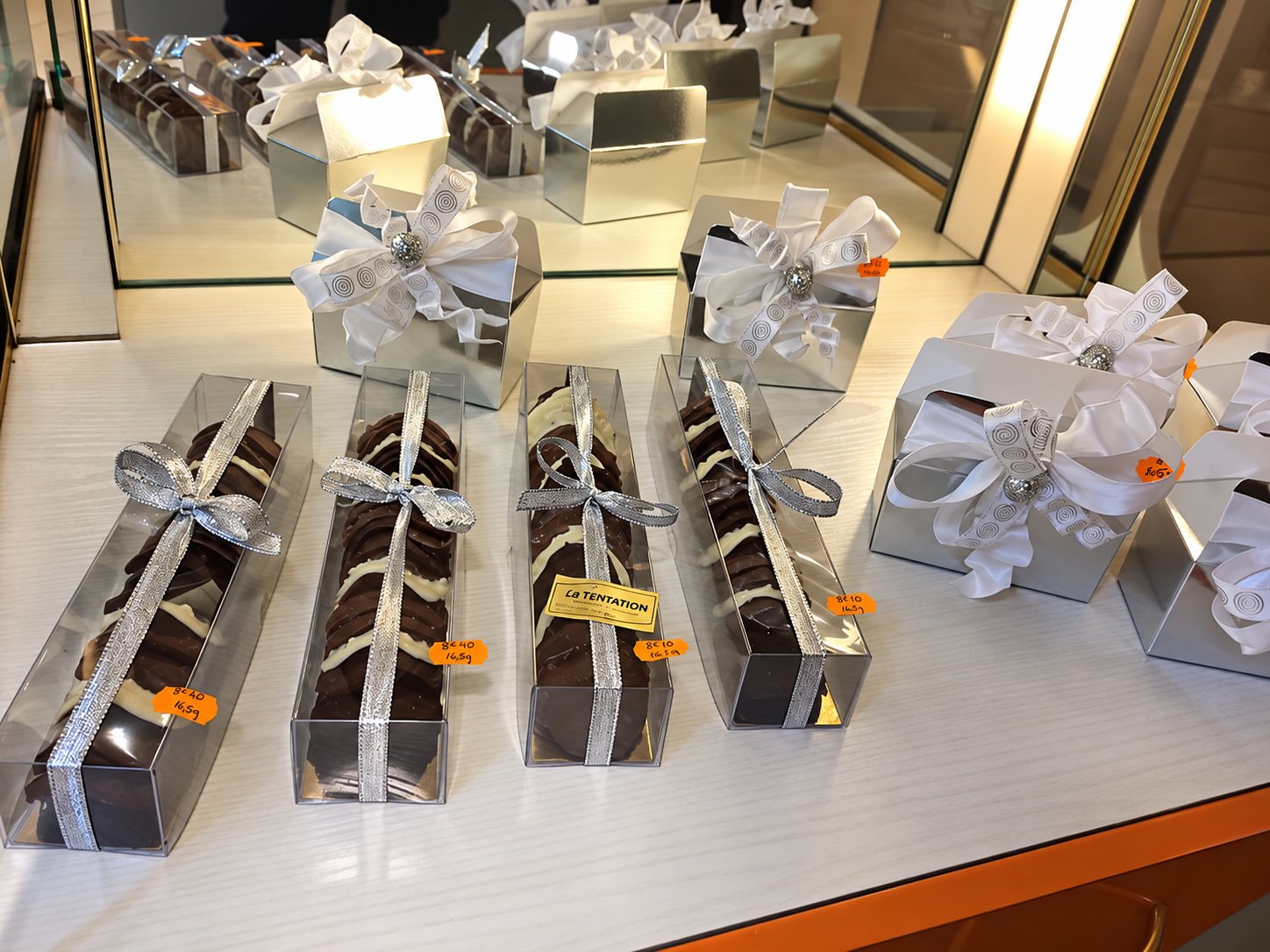 Vitrine de chocolatier : barquettes transparentes de chocolats noirs et blancs nouées de rubans argentés, et boîtes argentées.